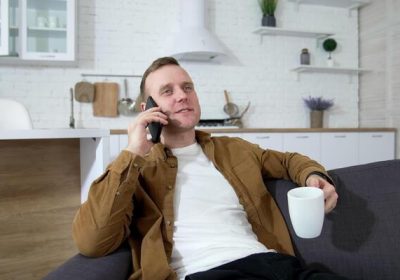 young-man-talking-the-phone-in-the-kitchen-guy-is-sitting-on-sofa-with-white-mug-and-speaking-on-a-mobile-phone-at-home-video
